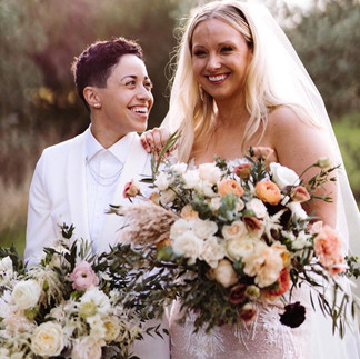 Two smiling people, one in a white suit and the other in a strapless gown, holding bouquets of vibrant flowers outdoors.