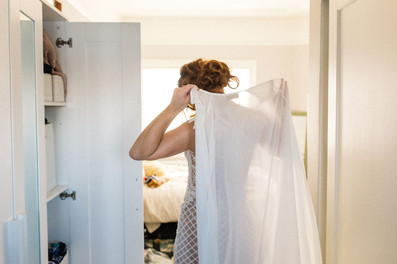 A woman, partially visible, holds a wedding cape in a softly lit bedroom. Open wardrobe and bed in the background suggest a calm, intimate moment.