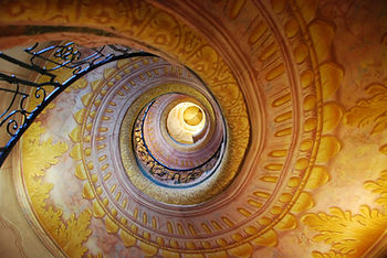 Ornate spiral staircase with intricate yellow and pink patterns, viewed from below. Black wrought iron railing enhances elegance. It is a visual paradox in that it is unclear if it is turning upwards or downwards.