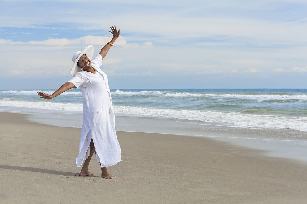 Woman in flowing white dress standing barefoot on a beach with arms open wide, facing the ocean and sky in an embodied gesture of openness and freedom.