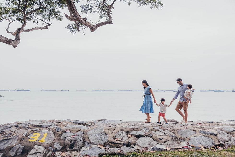 Family of four walking along a rocky shore, holding hands. Overcast sky, sea with distant ships. Tree branches frame the scene. Number 31 on rocks.