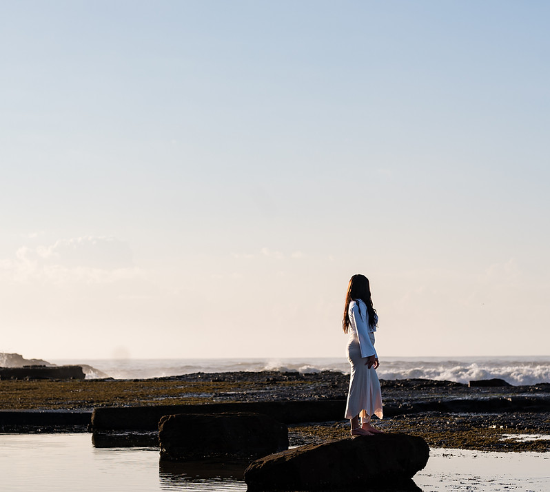 Stress Coach Steph Williams, The Rested Woman, standing on a beach rock looking out at the crashing waves wearing soft neutral coloured activewear and her long hair out.