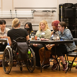 Photograph of several students seated at a table during the VSA Playwright Discovery Program. The workshop leader is seated in a wheelchair, instructing the group.