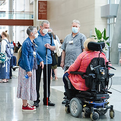 Photograph of four people interacting with each other in a lobby. Three are standing, one with a white cane. The fourth is seated in a motorized wheelchair.