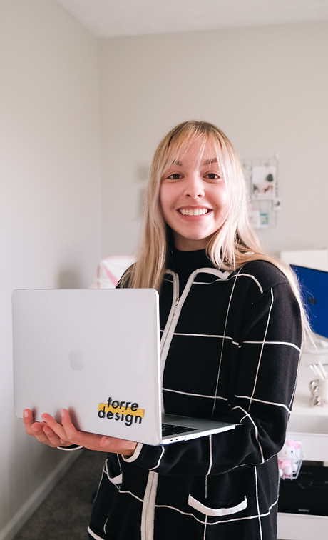 This is a headshot of Torre from Torre Design. She has long blonde hair and is smiling directly at the camera. She is holding an Apple laptop that features a sticker of the Torre Design logo. She is wearing a white and black grid pattern sweater. In the background, you can see a desk and photographs on the wall.