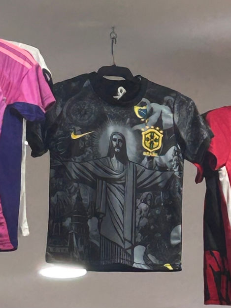 A football jersey printed with the image of Jesus Christ hanging beside colorful shirts. A contemporary reflection on faith, consumerism, and sacred imagery in modern culture.
