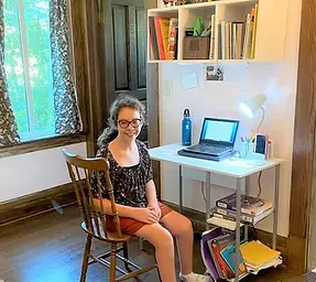 a white disabled young woman with glasses sits at a computer desk with her body turned towards the camera smiling