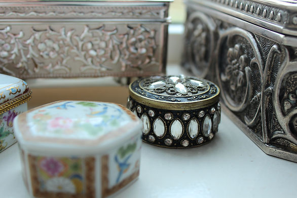 This is an image of a Marcasite trinket box placed on a windowsill. The box is made of a dark-colored metal, possibly silver or pewter, and it has a lid that is adorned with small, glittering marcasite gemstones. The box is round in shape and has a textured surface with intricate designs that create a vintage or antique look. The windowsill is made of wood and is painted white. Behind the box, there is a view of trees and foliage, suggesting that the windowsill is located in a natural outdoor setting. The overall image depicts a decorative and valuable trinket box placed in a charming and peaceful environment.