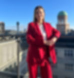 A woman dressed in red is standing on a balcony with buildings in the background.