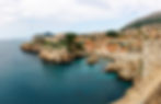 A panoramic coastal view of Dubrovnik, Croatia, featuring the Lovrijenac Fortress perched on a rocky cliff and the city's ter