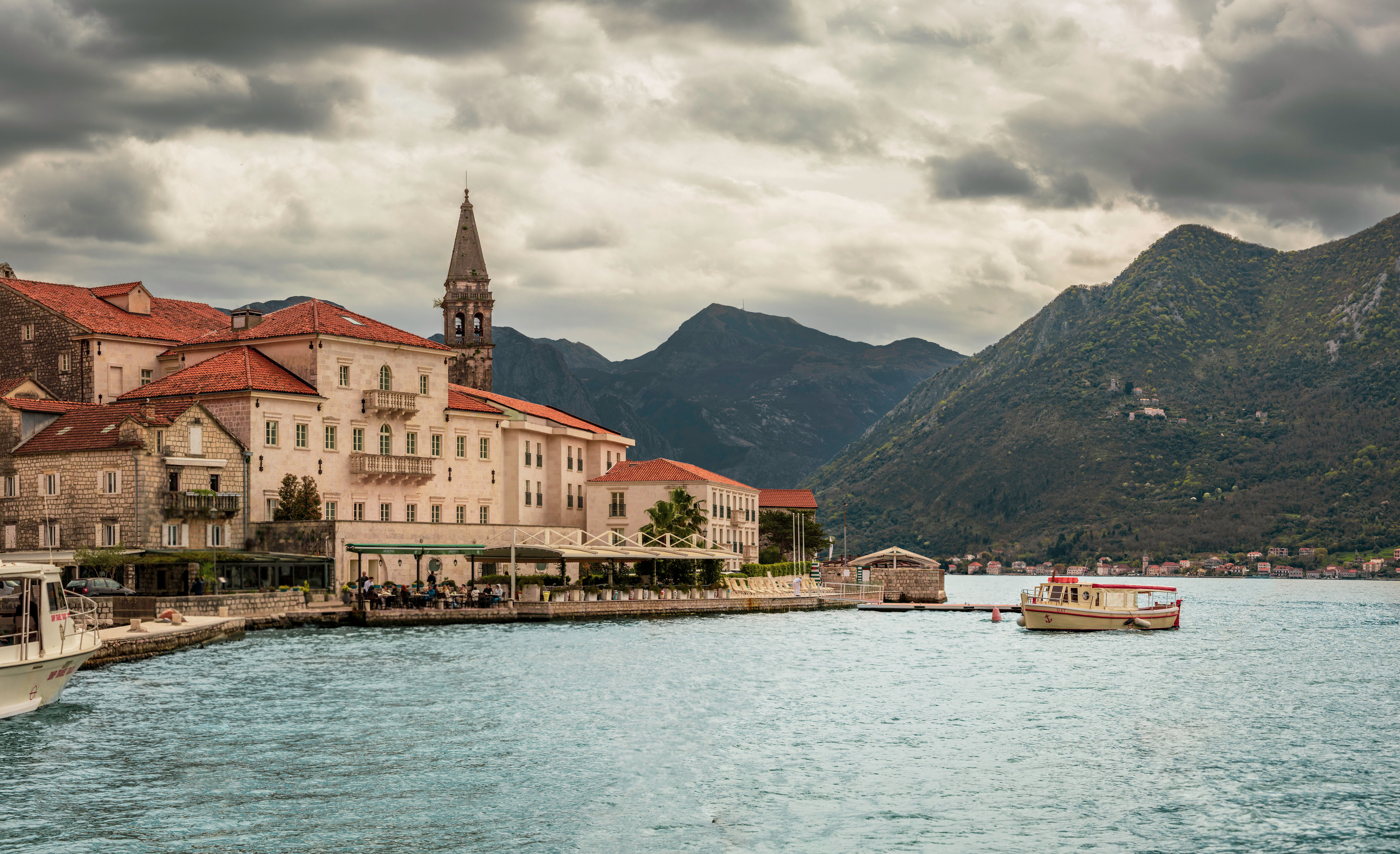 Perast, Montenegro, with red-roofed stone buildings, a bell tower, and boats floating on the Bay of Kotor, framed by dramatic