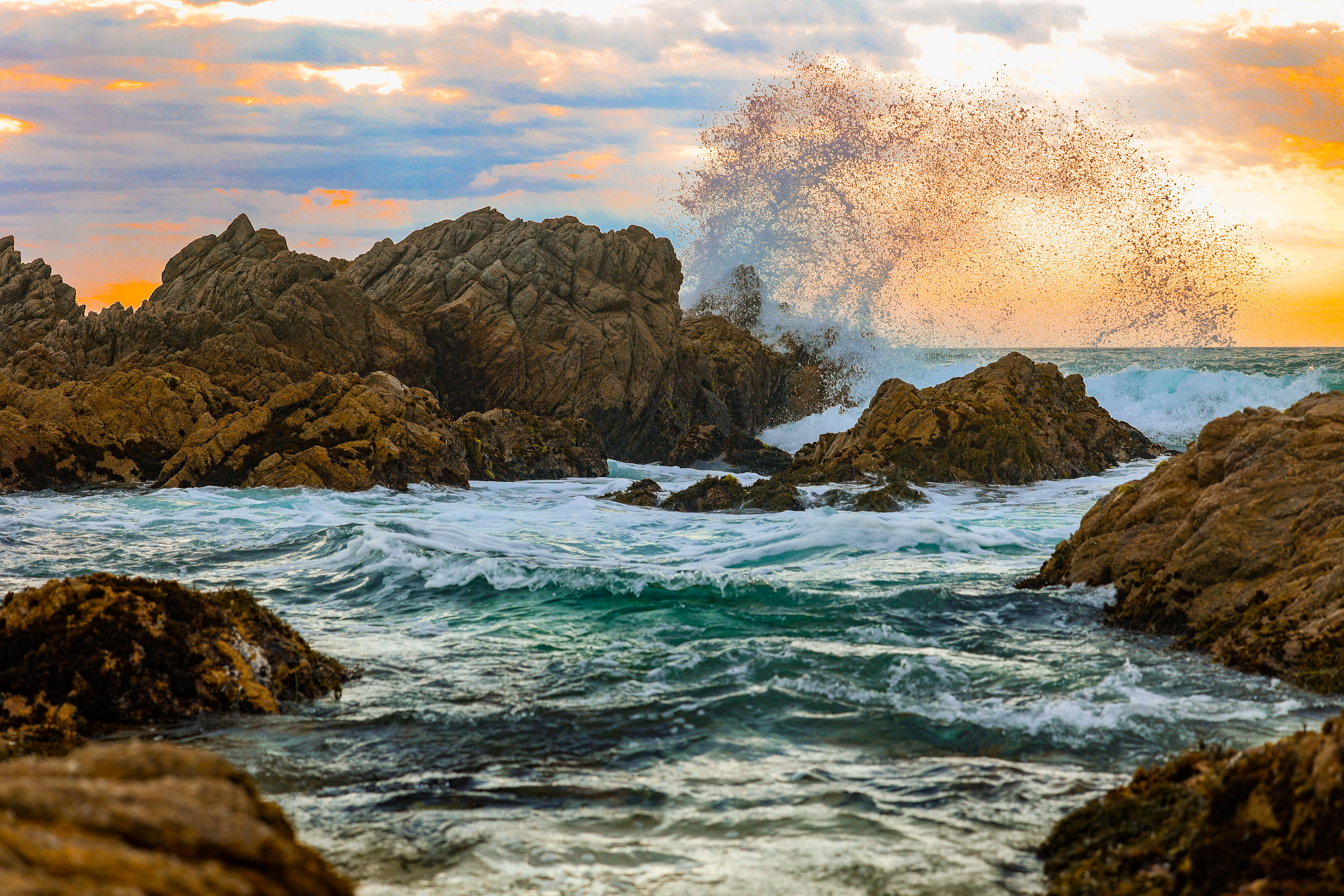 Waves crashing against rugged coastal rocks during a vibrant sunset in Carmel, California, with ocean spray caught mid air.