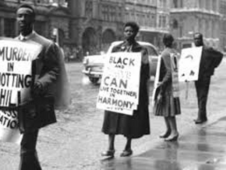 A black and white photograph depicts a protest march; several people carry signs, one reading "Murder in Notting Hill" and another proclaiming that "Black and White Can Live Together in Harmony".