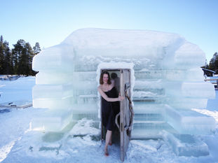 Woman exits an ice sauna wrapped in a towel, smiling. Snowy landscape with trees in the background under a clear blue sky.