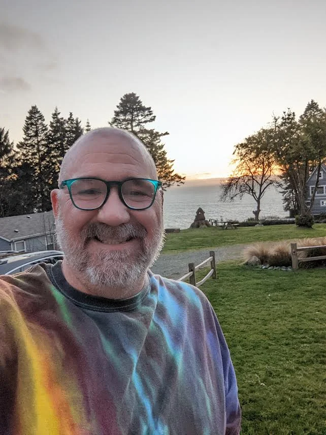Picture of Tom, a bald man with a grey beard, wearing blue glasses and a tie dye shirt. He is standing in front of a grassy area with a body of water and trees behind him.
