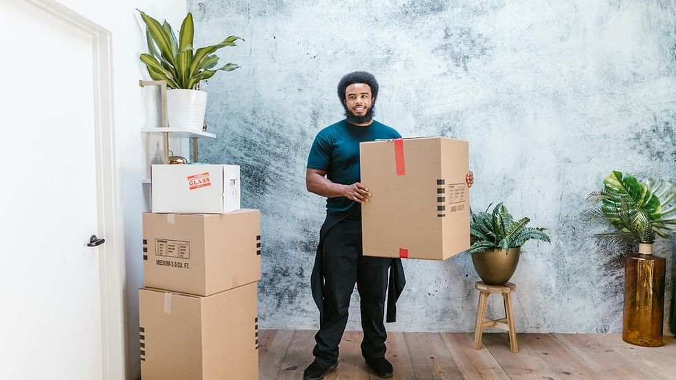 Afro-haired man wearing coveralls holding a cardboard box indoors surrounded by potted plants.