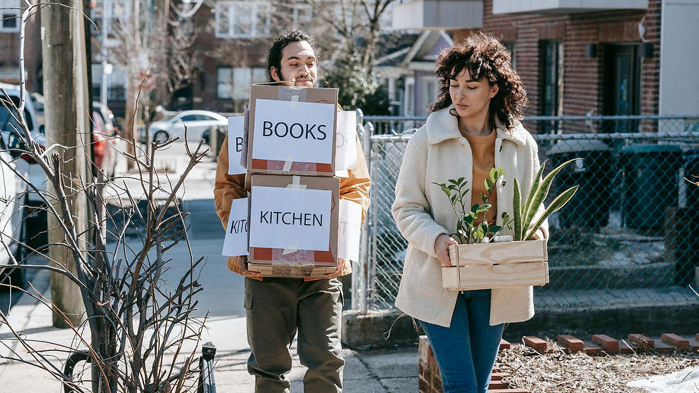 A couple carrying labeled boxes and plants, moving into a new urban neighborhood.
