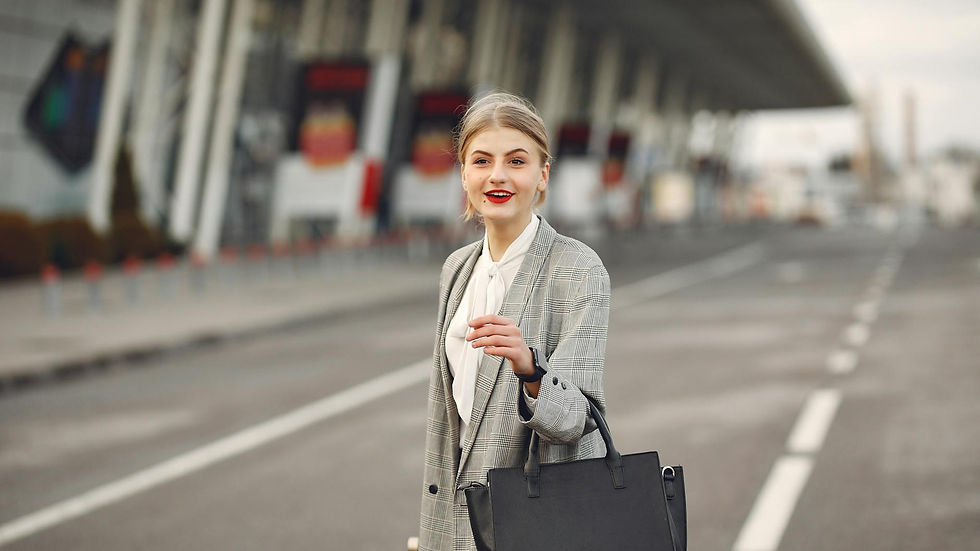 Positive female passenger wearing formal plaid suit standing on asphalt road to airport with handbag and suitcase while smiling looking at camera during business trip