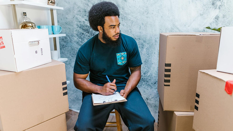 A focused male mover sits with a clipboard amid cardboard boxes, planning relocation tasks.