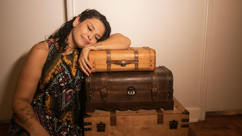 A woman with tattoos sits smiling, leaning on vintage suitcases in an indoor setting.