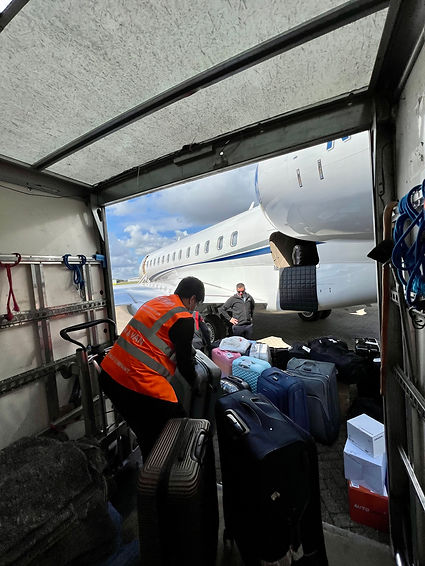 A VaiVan moving porter unloading suitcases for a airport transfer delivery service to a private jet client whilst the pilot of the chartered airplane watches the moving service take place by VaiVan