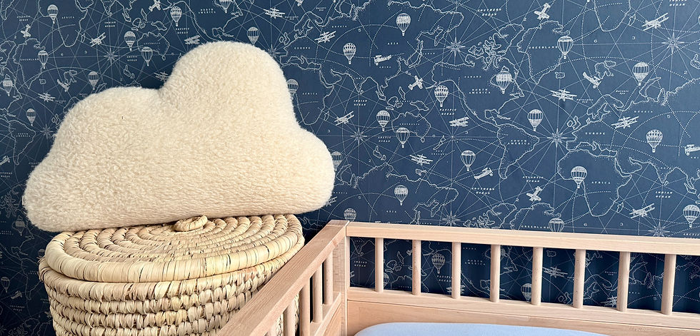 Knusse babykamer met wereldkaartbehang, een wieg van hout en een decoratief wolkvormig kussen van zachte teddy stof op een gevlochten mand.