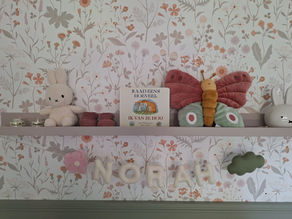 Babykamerdecoratie met een naam "NORAH" in vilten letters, omringd door knuffels, een kinderboek en een vlinder op een plank met bloemenbehang op de achtergrond.