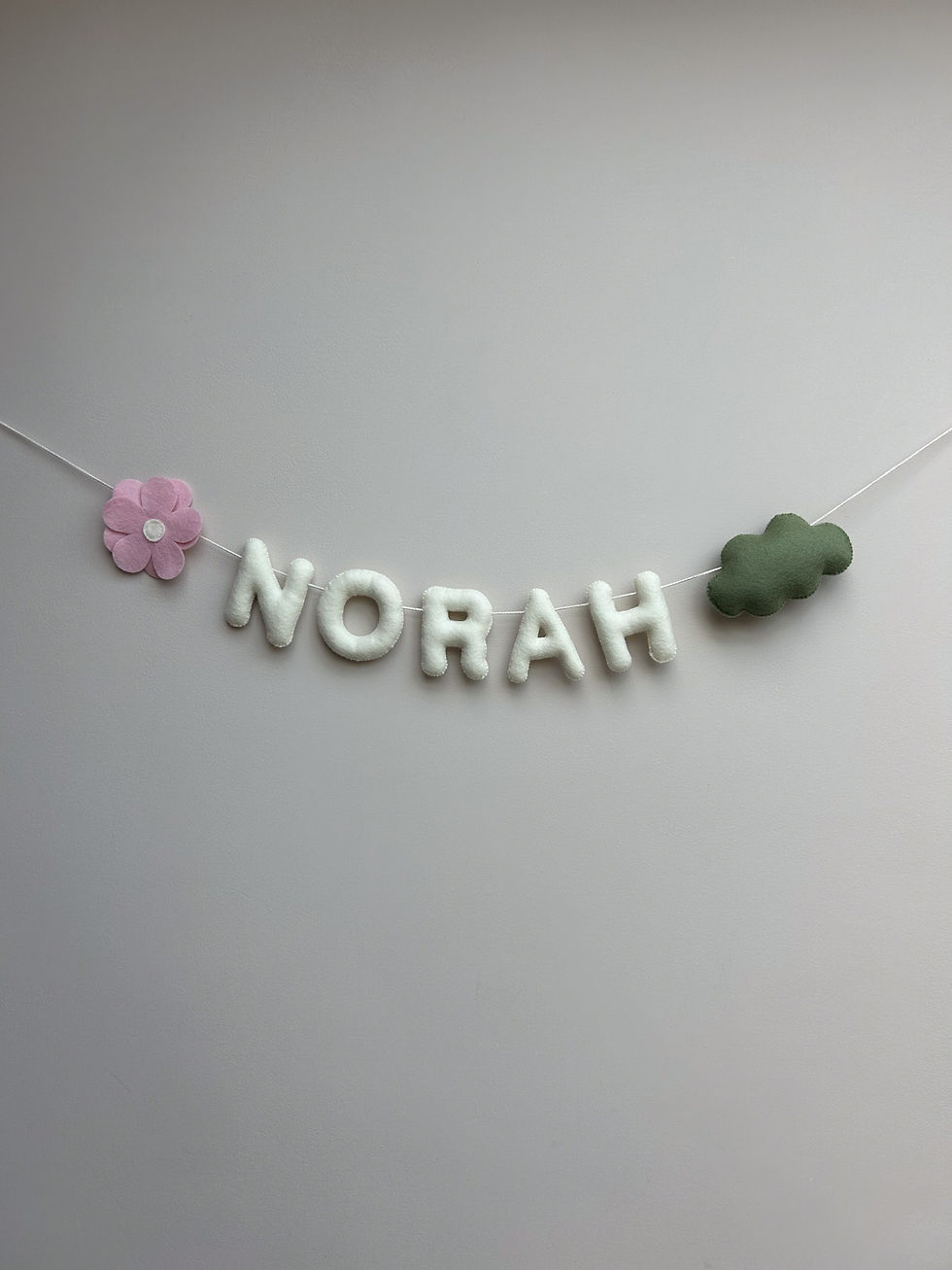 Handgemaakte vilten slinger met de naam "NORAH", versierd met een roze bloem en groene wolk, hangend tegen een effen grijze muur.