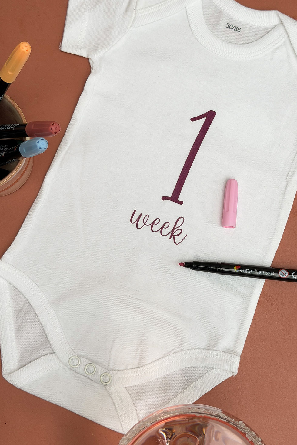 Thumbnail: Close-up van een romper met “1 week” uit een mijlpaalset, beschilderd tijdens een babyshower workshop.