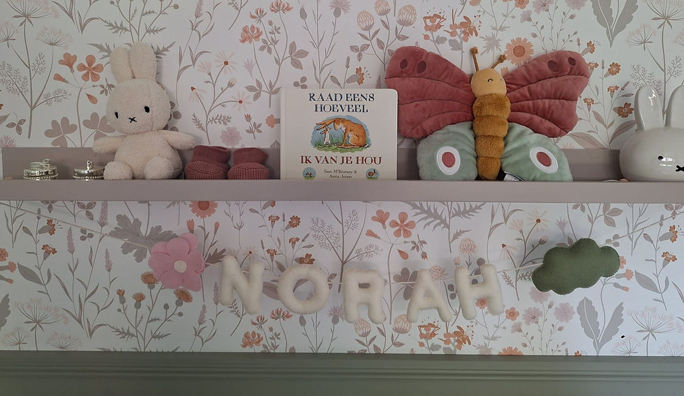Babykamerdecoratie met een naam "NORAH" in vilten letters, omringd door knuffels, een kinderboek en een vlinder op een plank met bloemenbehang op de achtergrond.
