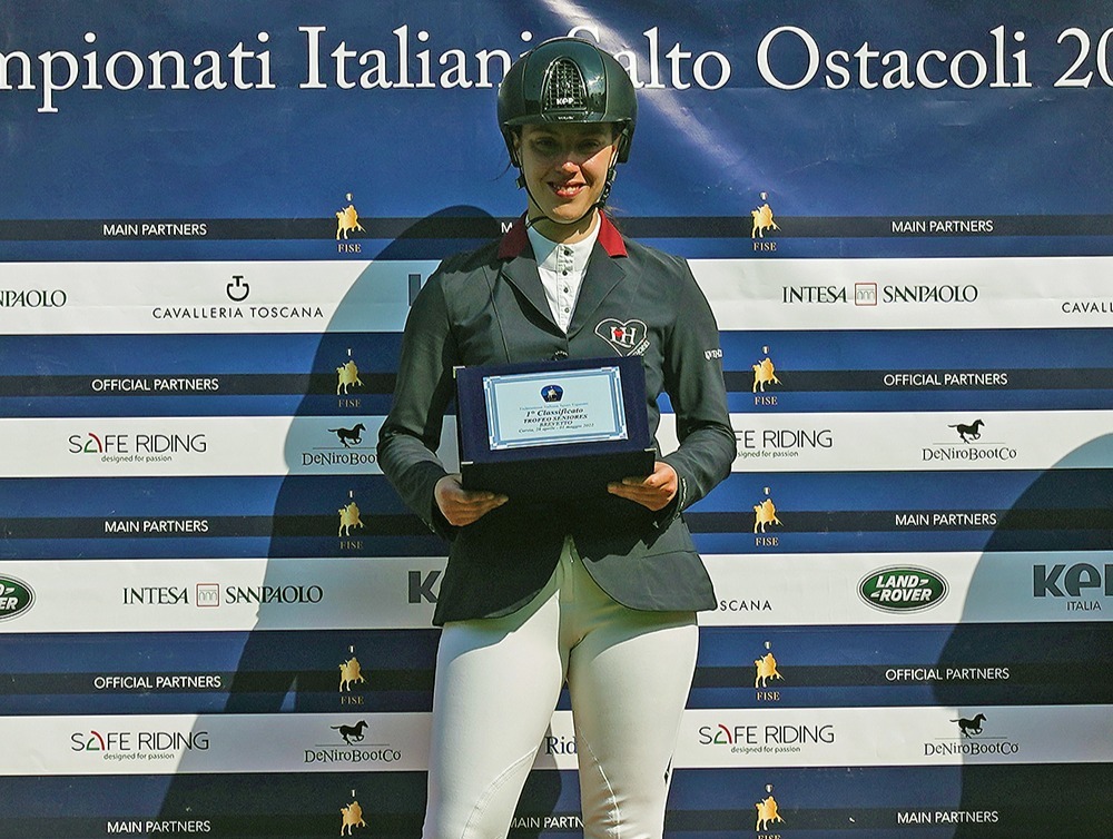 Sarah Tintori ĆØ in piedi sul podio, al centro della scena. Indossa la giacca LoveHorses confezionata da Sarm Hippique in collaborazione con LoveHorses  In mano ha un targa.  Sarah sembra molto felice e orgogliosa del suo successo, con un sorriso luminoso sul viso e gli occhi che brillano di soddisfazione.