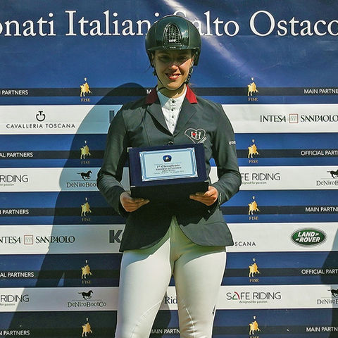 Sarah Tintori è in piedi sul podio, al centro della scena. Indossa la giacca LoveHorses confezionata da Sarm Hippique in collaborazione con LoveHorses In mano ha un targa. Sarah sembra molto felice e orgogliosa del suo successo, con un sorriso luminoso sul viso e gli occhi che brillano di soddisfazione.