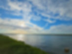 Sunny river landscape under a bright blue sky with wispy clouds.