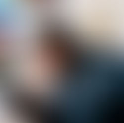 Foto em plano médio e ângulo diagonal de um homem com barba e coque no cabelo, usando uma camisa azul, sentado com o corpo inclinado para a frente. Ele olha com expressão de desânimo para o smartphone que segura na mão esquerda, enquanto a direita apoia a cabeça e coça o rosto. Ao fundo, está uma parede com diversas fotos em tamanhos e cores diferentes desfocadas.