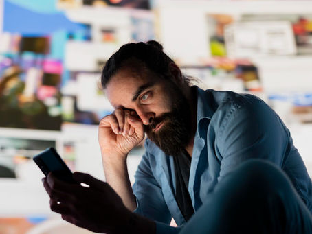 Foto em plano médio e ângulo diagonal de um homem com barba e coque no cabelo, usando uma camisa azul, sentado com o corpo inclinado para a frente. Ele olha com expressão de desânimo para o smartphone que segura na mão esquerda, enquanto a direita apoia a cabeça e coça o rosto. Ao fundo, está uma parede com diversas fotos em tamanhos e cores diferentes desfocadas.