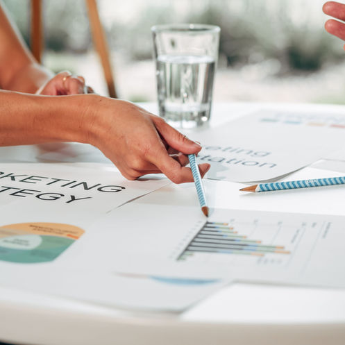 Uma equipe reunida em torno de uma mesa analisa gráficos e documentos de estratégia de marketing. Uma pessoa aponta para um gráfico com um lápis enquanto outra gesticula durante a conversa. Sobre a mesa há relatórios impressos, um gráfico circular, lápis e um copo de água, sugerindo uma reunião de planejamento ou alinhamento entre marketing e vendas.