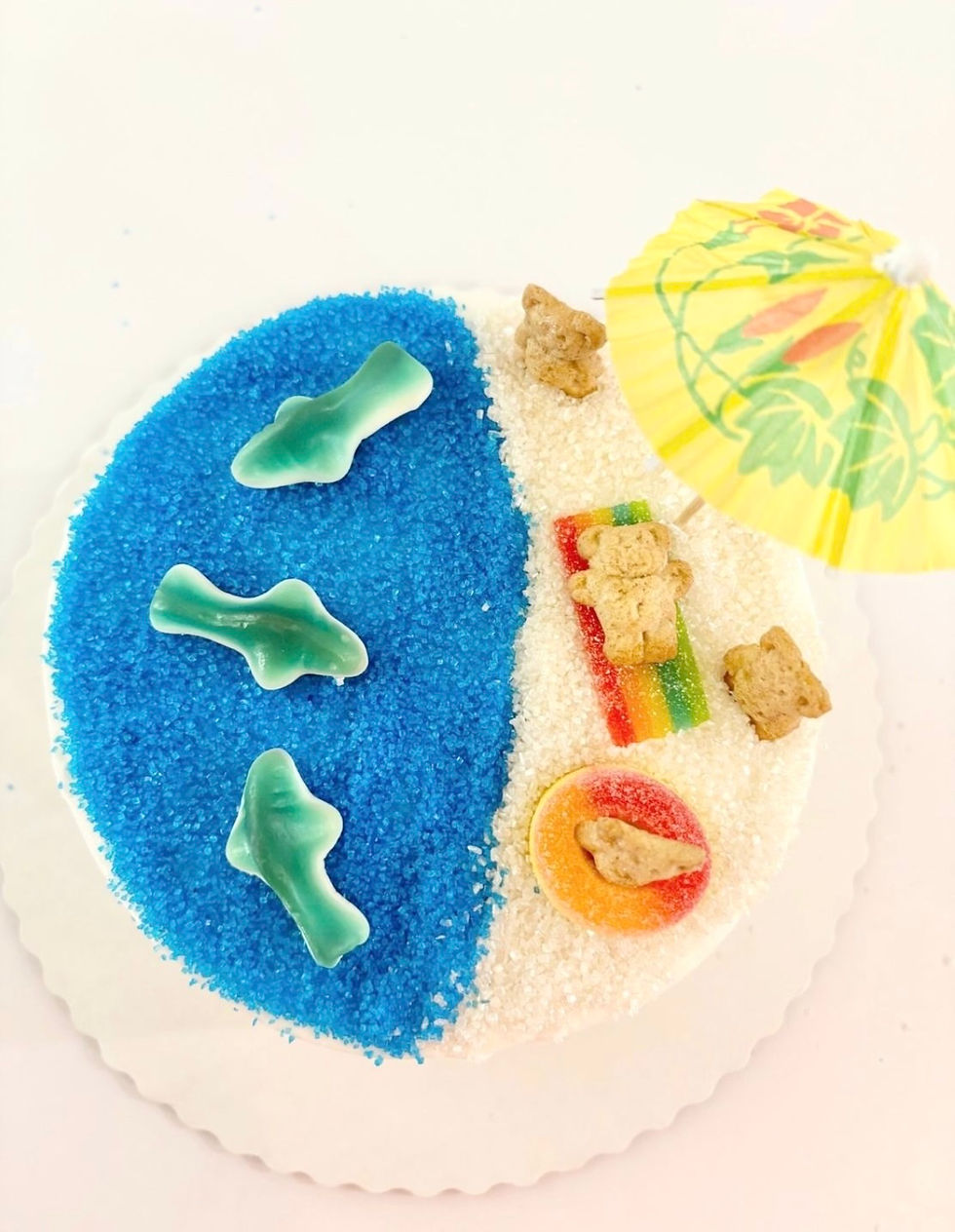 A summer beach-themed cake. Blue and white sugar crystals act as the water and the sand. Blue gummy sharks sit in the water, while Teddy Graham bears relax under a mini umbrella on the beach.