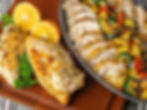Sliced grilled chicken with vegetables and herbs in a black dish, next to whole chicken breasts, oranges, and parsley on a wooden board.