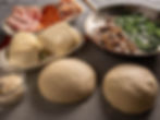 Pizza ingredients on a table: two dough balls, sliced meats, cheese, a pan with mushrooms and spinach, and a bowl of grated cheese.