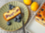 Lemon blueberry loaf slice on a green plate with fresh blueberries, fork, and whole lemons on a textured tablecloth background.