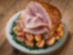 Slice of glazed ham on a platter with roasted potatoes and greens on a wooden table. The ham looks juicy and inviting, set in a rustic mood.