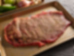Seasoned raw beef on a baking sheet with spices, surrounded by green peppers, onions, and garlic. Set on a black and white checkered cloth.