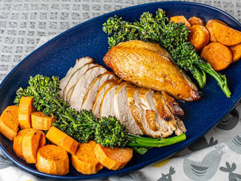 Roast turkey breast sliced on a blue platter with broccolini and roasted sweet potatoes. Background shows a patterned tablecloth.