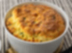 Golden brown souffle in a white ramekin on a striped surface, featuring a fluffy, textured top with hints of green herbs.