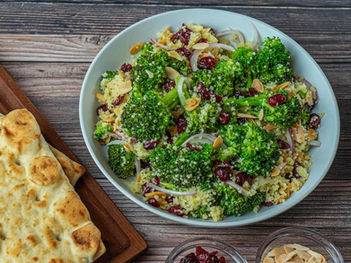 Bowl of broccoli couscous with almonds and cranberries. Flatbread on a wooden table nearby. Rustic and colorful presentation.