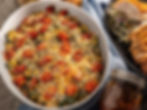 Baked veggie dish topped with cheese and tomatoes in a white bowl. Sliced chicken with coleslaw on a black plate nearby. Cozy dining setup.