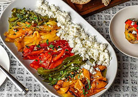 Roasted peppers with feta on a platter, surrounded by sliced bread on a board. Plates with forks are set on a patterned tablecloth.