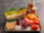 Spices, apricots, tomatoes, garlic, and onions on a tray with jars of preserves. Packs labeled coriander and cumin. Rustic kitchen setting.