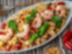 Oval plate with shrimp orzo salad garnished with basil leaves on a striped surface. Nearby are bowls of tomatoes and pine nuts.
