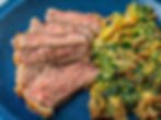 Steak slices and greens on a blue plate, with salt and pepper bowls. A wooden board holds more steak. Striped cloth in background.